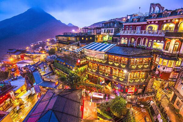 Beleuchtete Häuser und Gassen in Jiufen in Taiwan bei Abendlicht mit Bergkulisse im Hintergrund