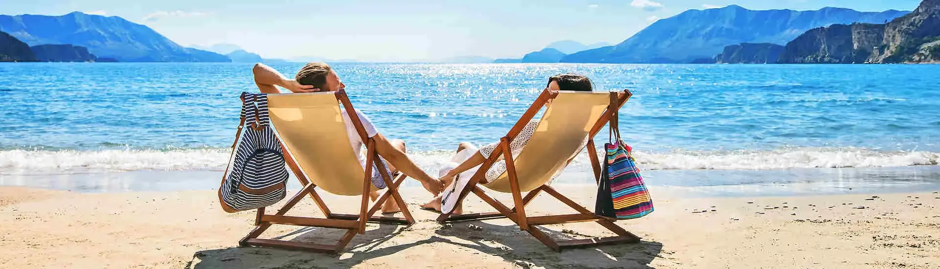 Zwei Personen entspannen auf Sonnenliegen am Strand mit Blick auf Meer und Berge, Symbol für weltweit kombinierbare Urlaubsziele