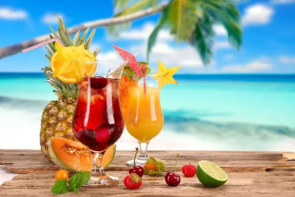 Zwei bunte Cocktails mit tropischen Früchten auf einem Holztisch vor einem weißen Sandstrand und türkisblauem Meer auf den Malediven