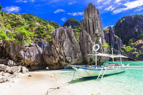 Traditionelles Auslegerboot an einem weißen Sandstrand vor markanten Kalksteinfelsen in Palawan auf den Philippinen.