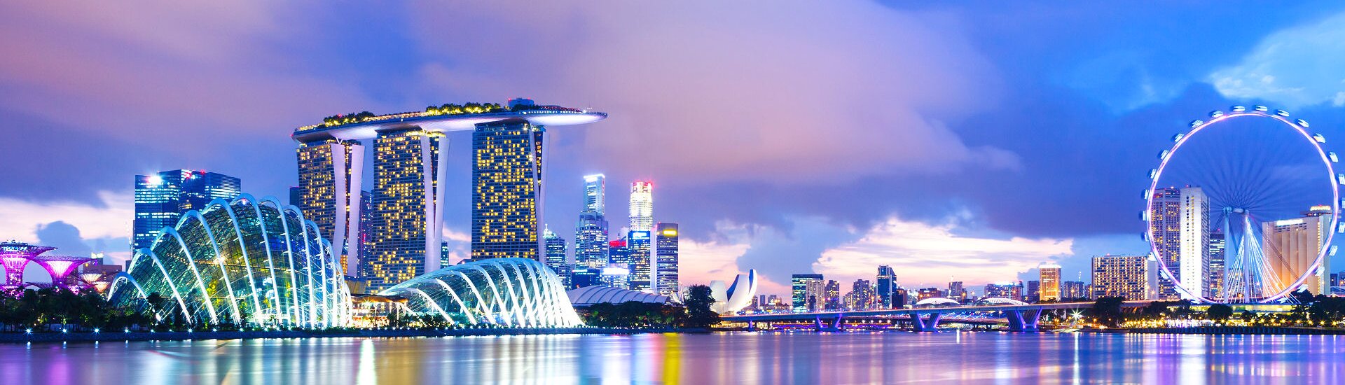 Marina Bay in Singapur mit dem Marina Bay Sands Hotel, den Gewächshäusern der Gardens by the Bay und dem Singapore Flyer bei Abendbeleuchtung.