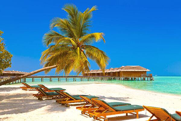 Sonnenliegen unter einer Palme am weißen Sandstrand mit Blick auf Overwater-Villen und türkisfarbenes Meer auf den Malediven