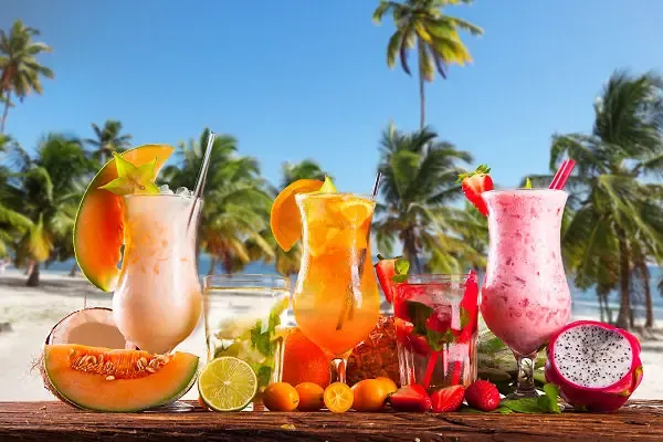 Zwei bunte Cocktails mit Früchten und Strohhalmen auf einem Holztisch vor einem tropischen Strand mit Palmen