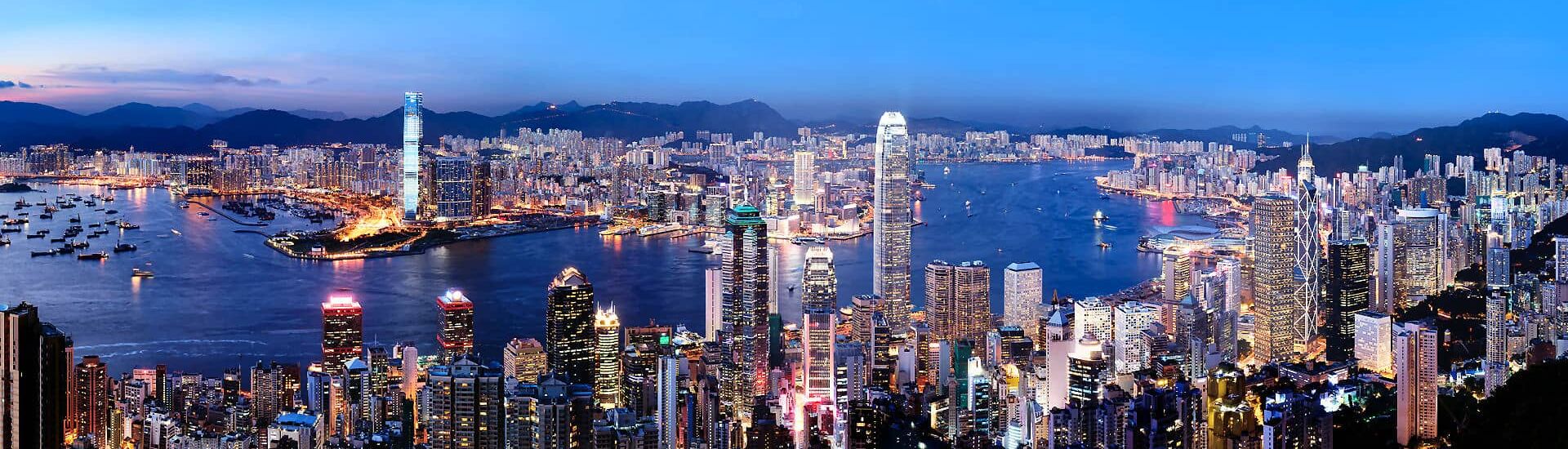 Panoramablick auf die beleuchtete Skyline von Hongkong mit Victoria Harbour, Wolkenkratzern und zahlreichen Schiffen bei Nacht.
