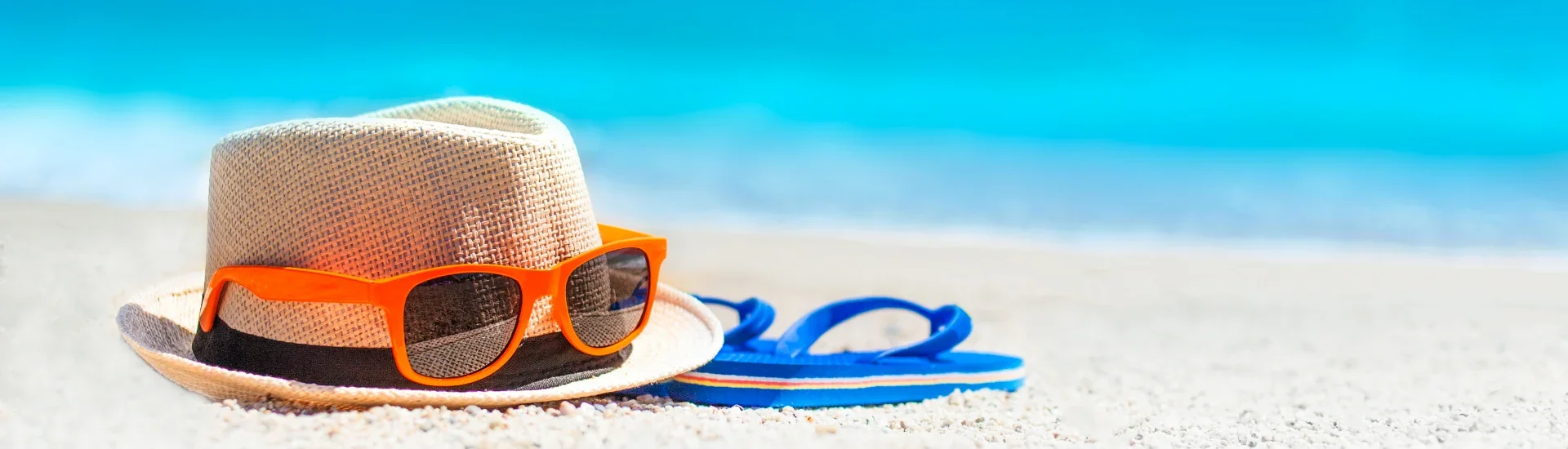 Sonnenhut, orangefarbene Sonnenbrille und blaue Flipflops liegen am Strand vor türkisblauem Meer