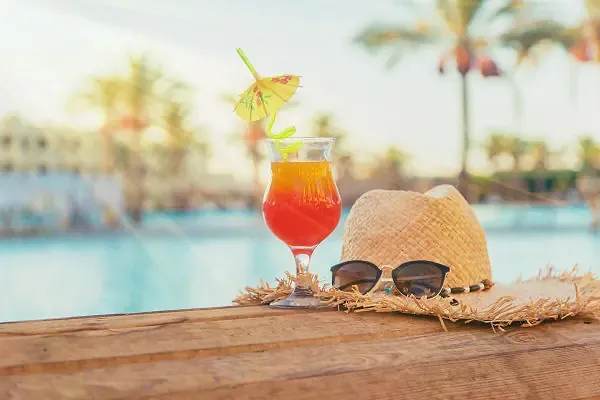 Zwei bunte Cocktails mit Früchten und Strohhalmen auf einem Holztisch vor einem tropischen Strand mit Palmen