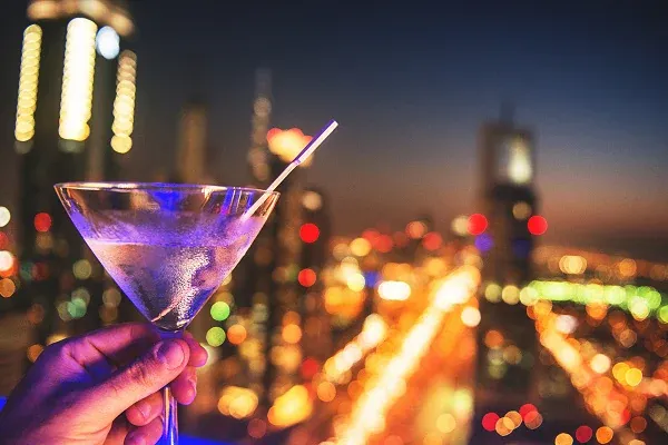 Hand mit Cocktailglas in einer Rooftop-Bar über der beleuchteten Skyline von Dubai bei Nacht