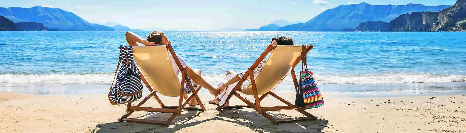Paar entspannt auf Sonnenliegen am Strand mit Meer und Bergen – Kombireisen