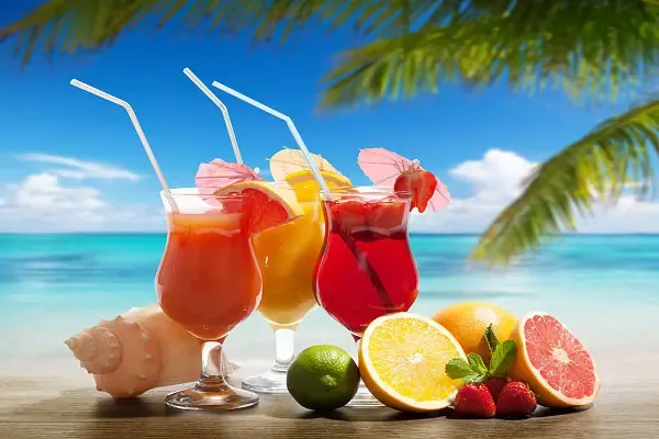 Zwei bunte Cocktails mit Früchten und Strohhalmen auf einem Holztisch vor einem tropischen Strand mit Palmen
