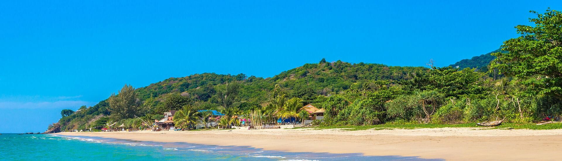 Langer Sandstrand am Klong Jark Beach auf Koh Lanta mit Palmen, kleinen Strandhäusern und grünem Hügel im Hintergrund
