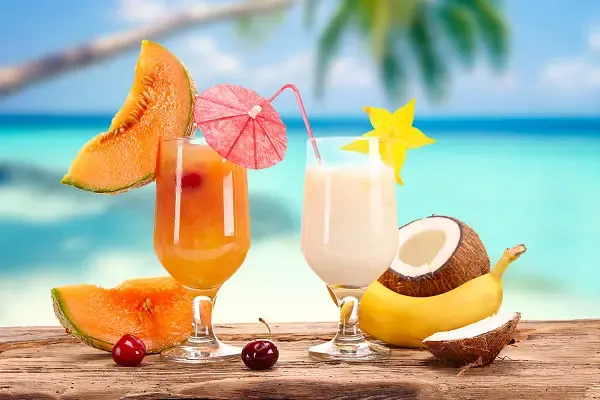 Zwei farbenfrohe Cocktails mit tropischen Früchten auf einem Holztisch vor einem Strand mit Palmen und blauem Meer