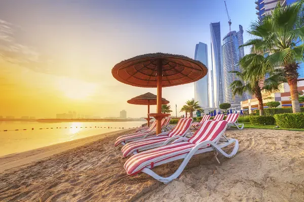 Strandliegen und Sonnenschirm am Corniche Beach in Abu Dhabi vor moderner Skyline bei Sonnenaufgang