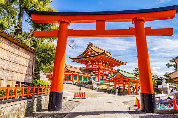 Rotes Torii-Tor am Fushimi Inari-Taisha Schrein in Kyoto, Japan, mit traditionellem Tempelgebäude im Hintergrund.